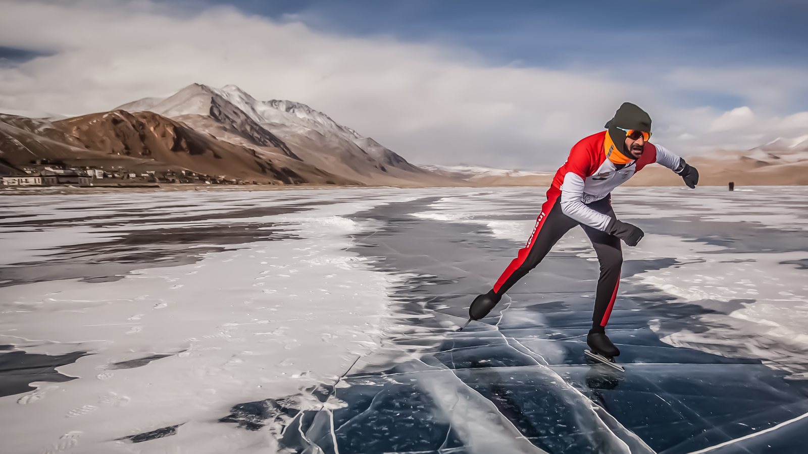 Vishwaraj skating across blue black ice on a Ladakh lake, village and snow peaks behind